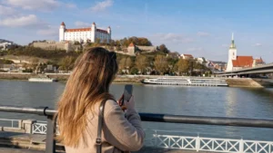 Tourist using a smartphone beside the Danube promenade with Bratislava Castle in view.