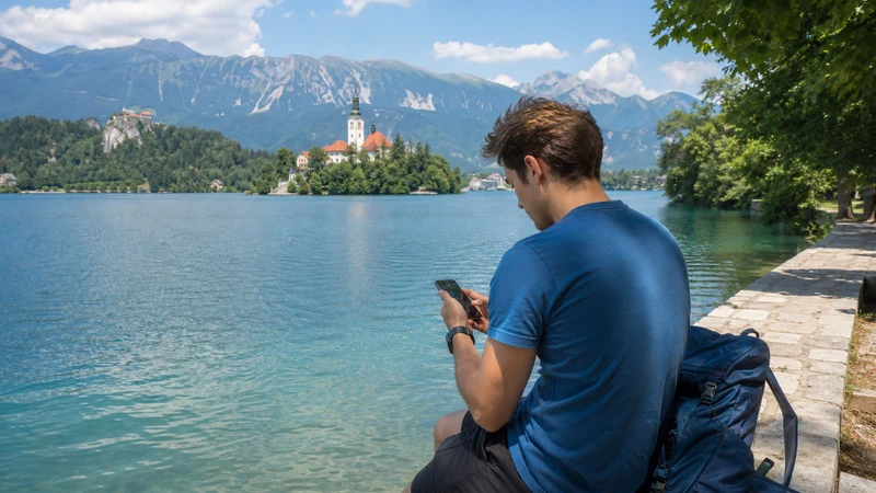 Traveller using mobile data beside Lake Bled and the Julian Alps.