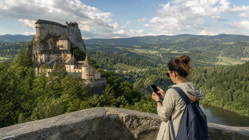 Visitor checking a smartphone at a viewpoint overlooking Orava Castle and its valley.