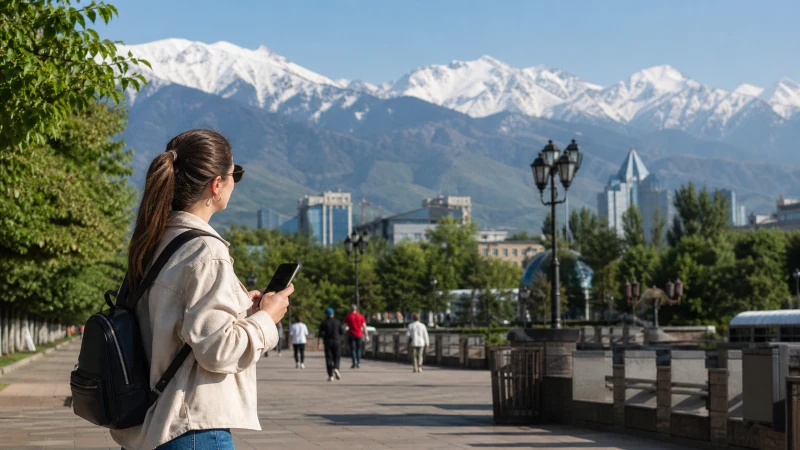 Tourist using a smartphone on a central Almaty boulevard with mountains behind.