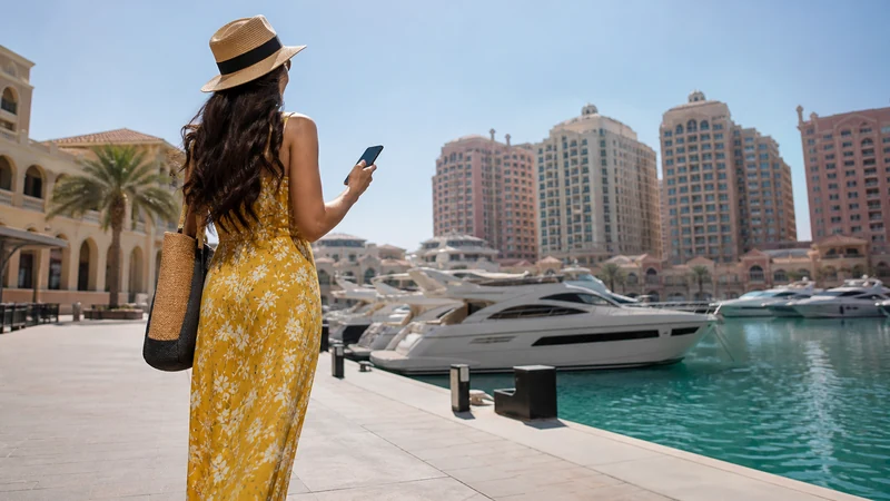 Traveller using a smartphone in visitor beside yachts and pastel buildings at the Pearl-Qatar